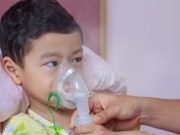 Close-up nurse’s hand helping sick child hold nasal mask with respiratory problem in hospital room. Asian boy patient inhalation therapy by the mask of inhaler with bronchodilator’s soft stream smoke