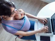 Pretty young mother with her baby in sling working with laptop at home.