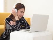 Young Asian student using laptop computer and sitting on floor in living room while listening and watching online coursework with electronic earphones or headphone. Internet E-Learning concept